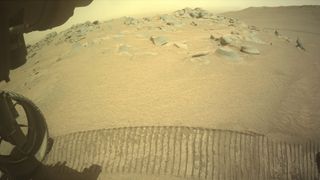 closeup image taken by a mars rover, showing one of its wheels and a track, with rocks and red dirt in the distance 