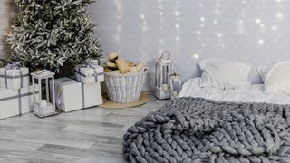 Image shows a spare room mattress placed on the floor and covered with white linens and pillows while a frosted green christmas tree sits in the corner surrounded by brightly wrapped presents 