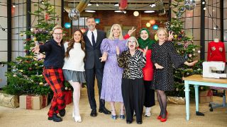 Judges Patrick Grant, Esme Young, and host Sara Pascoe posing with this year&#039;s celebrity contestants and flanked by a festive backdrop of bauble-adorned Christmas trees for The Great British Sewing Bee Christmas Special 2024.