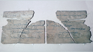 A fragmented wooden tablet with Latin writing on it in charcoal ink on display against a neutral background