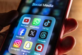 A man holds a smartphone iPhone screen showing various social media apps including YouTube, TikTok, Facebook, Threads, Instagram and X