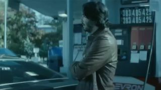 Keanu Reeves in profile, wearing a brown leather jacket, standing next to a gas station pump in John Wick