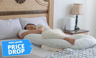 A woman with dark hair and wearing light-coloured bed clothes sleeps on her side while hugging a full body pillow