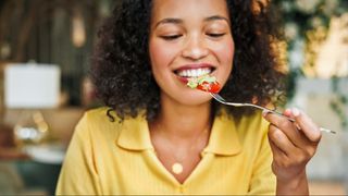 A woman with brown curly hair wearing a yellow top smiling as she lifts a fork of food to her mouth