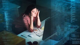 A shot of a woman sat at her desk in a dimly lit office, with her eyes closed and a stressed expression on her face, her hands raised to massage her temples. In the foreground, blue code is rising to either side of the frame to indicate complexity in the task that her unseen screen is showing