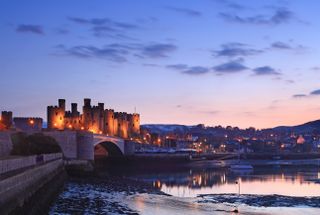 A classic dusk shot of Conwy Castle in Wales, with bridge leading in and town lights glowing in the background