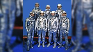 seven men stand in shiny silver spacesuits four stand on the front row and three at the back.