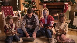 Young parents and their kids sitting on the floor on New Year&#039;s eve and using wireless technology.