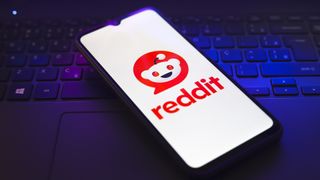 The Reddit logo appears on the screen of a smartphone that rests on top of a laptop keyboard.