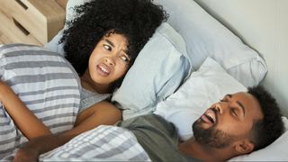 A couple in bed where the man is snoring and the woman is looking at him frustrated