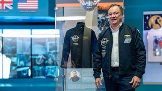 a smiling older man in a blue jacket stands next to a blue flight suit inside an air and space museum