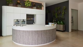 A curved kitchen island with wood cladding and white units behind with plants on top