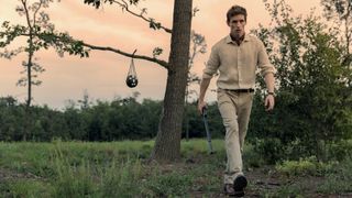 Eddie Redmayne as the Jackal walking away from a ball in a net hanging on a tree with a gun in his hand.