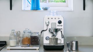 a photograph of the black smeg mini pro espresso machine with from a kitchen environment with a milk jug and tamper and milk bottles