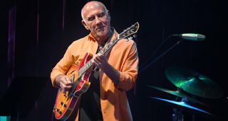 Larry Carlton wears an orange shirt and plays a cherry sunburst semi-hollow electric guitar onstage in Japan