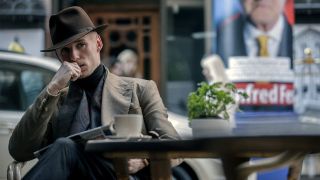 The stylishly dressed Jackal (Eddie Redmayne) dons a fedora and ponders his next move at a outdoor cafe in the British TV thriller The Day of the Jackal 