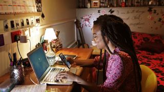 Student sat at a desk with a laptop in a dormitory looking at a mobile phone