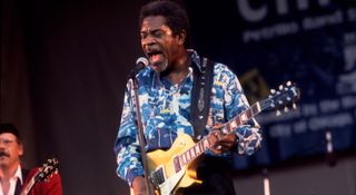 Luther Allison rocks the crowd at the 12th Chicago Blues Festival 1995. He wears a blue-and-white pattern shirt and plays a Gibson Les Paul Standard Gold Top.