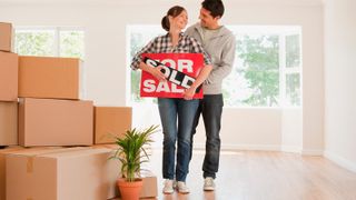 A couple holding up a sign saying FOR SALE and SOLD in an empty house