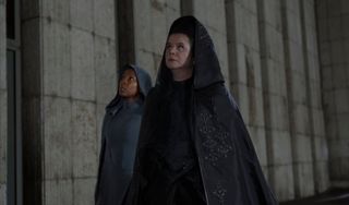 Two cloaked women stand looking up at a stone fortress