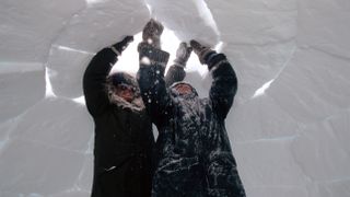 Two Inuit people wearing black coats work above their heads on their igloo