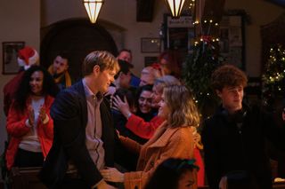 A scene in the church in Shipton Abbott, where Humphrey (Kris Marshall) and Martha (Sally Bretton) are moving in for an embrace. Jaiden (Austin Taylor) is standing next to Martha, with a set of headphones hanging around his neck, looking in the other direction. The church is decorated for Christmas and in the background we can also see other characters embracing each other warmly including Anne (Barbara Flynn), Margo (Felicity Montagu), Zoe (Melina Sinadinou) and Chief Superintendent Woods (Jade Harrison).