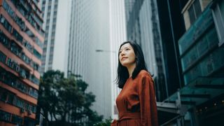 A happy and thoughtful-looking businesswoman is standing in a downtown business area surrounded by tall glass buildings