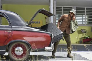 DI Mervin Wilson (Don Gilet) is at the airport, wearing sunglasses and a sunhat, using one hand to lift a large wheelie suitcase out of the car while the other hand holds his mobile phone to his ear. He looks annoyed and stressed.