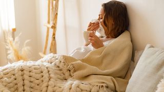 A woman with dark hair sits up in bed blowing her nose because she has a cold