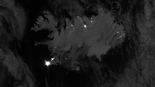 a dark view from above Earth shows an icy landmass speckled on its edges with bright dots of light, some clustered as well. 