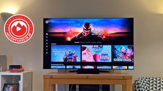 A 65-inch Samsung TV on a wooden stand in a living room. On the screen is the homepage of Sony Pictures Core from the PS5, with a big banner for the Venom: The Last Dance movie.