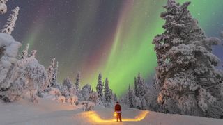 christmas northern lights. Green ribbons of light in the sky and snow on the ground. A man dressed as Santa holds a lantern. 