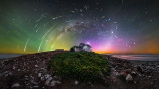 A photograph of the northern lights with meteors and the milky way over a house on a rocky shore