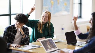 Some teenage students are sitting round a table looking towards a teacher, who is out of shot. A girl with red hair and a green hoodie is smiling with her hand raised. Another student who is slightly out of focus in the foreground also has her hand raised and is working on a laptop.