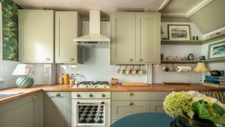 A kitchen with sage-green shaker cabinets, a white SMEG oven, wooden countertops, open shelves displaying decor and mugs, and floral accents 