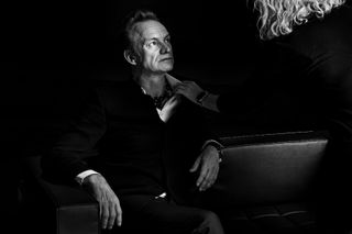 a black and white portrait of the singer Sting sat on a chair looking to the right