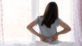 a photo of a woman sat on her bed, holding her lower back 