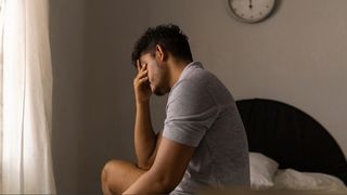 A man wearing a grey tshirt sitting on bed of bed in beige room with his head in his hands as he struggles to sleep with anxiety and depression.