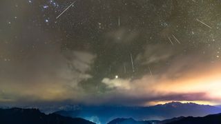 A breathtaking night sky filled with streaks of light from the Geminid meteor shower, set against a backdrop of twinkling stars and partially cloudy skies. Below, rugged mountain peaks are silhouetted, with a faint glow of city lights casting a soft illumination. The horizon features vibrant hues of orange and purple, blending into the dark, starry expanse above.