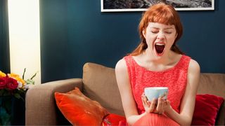 A woman with red hair and a fringe, wearing a red dress sitting with a tea cup in her hands yawning as she survives off four hours sleep