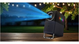 The projector emits a beam of blue light into the top corner. It is on a wooden table outdoors, with a background of green trees and fairy lights. It is night time.