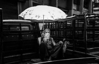 Black-and-white image of a man holding an umbrella while sitting in the back of a van, wearing a COVID mask.