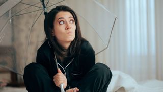 woman wearing black pyjamas sat on bed holding clear plastic umbrella and looking concerned