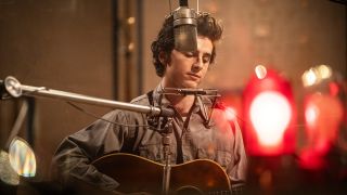 Timothée Chalamet as Bob Dylan in A Complete Unknown. He is playing guitar in a recording booth. There is a harmonica attached to him and a microphone near his face.