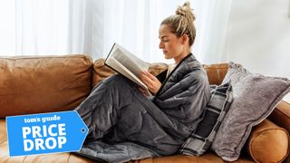 A woman curled up on the sofa reading a book and wrapped in the Layla weighted blanket