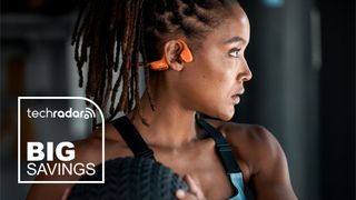 Lifestyle image showing woman working out with medicine ball while wearing Shokz OpenRun Pro 2 headphones in orange color option. A TechRadar &#039;Big Savings&#039; badge appears overlaid at the bottom left.