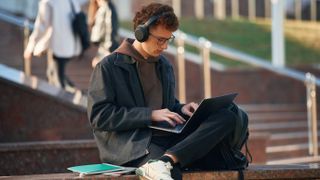 A male student using a laptop near a college or university, sitting outside