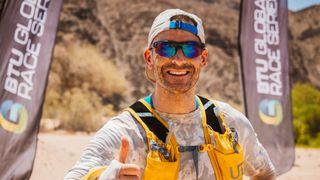 Ultra runner Jon Shield stands in a desert landscape in front of a race arch, wearing sunglasses, a cap and a hydration vest