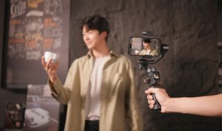 A man stands in a coffee shop holding a mug while another hand holding a Hohem iSteady M7 gimbal to record.