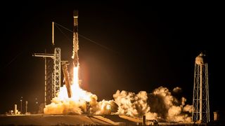 a white rocket launches at night above a bright cone of fire and smoke